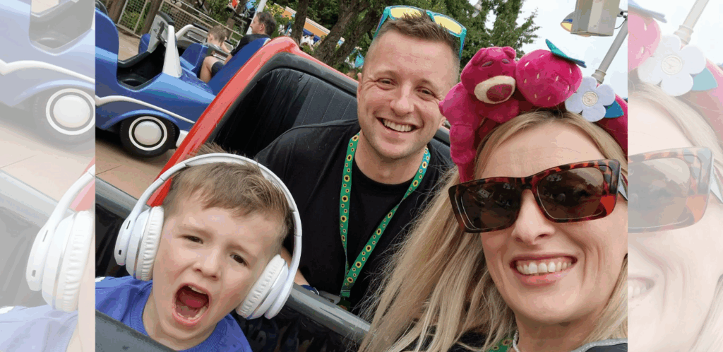 A smiling woman in sunglasses, a man, and a young boy wearing large headphones pose for a selfie while sitting in a theme park ride car. The background shows another blue ride car and some greenery.