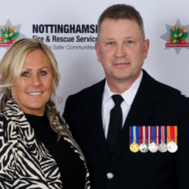 A man in a formal uniform with medals stands next to a smiling woman in a patterned jacket, both posing in front of a Nottinghamshire Fire & Rescue Service backdrop.