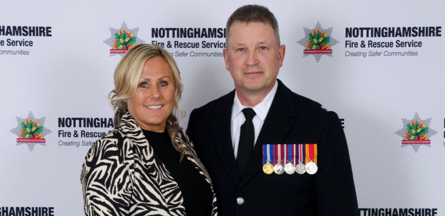 A man in a formal uniform with medals stands next to a smiling woman in a patterned jacket, both posing in front of a Nottinghamshire Fire & Rescue Service backdrop.