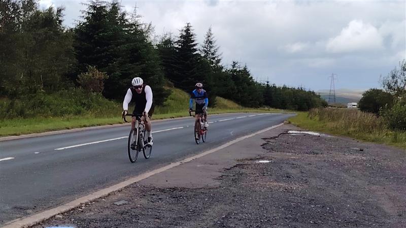 Two cyclists ride on a rural road bordered by trees and grass under a cloudy sky. The cyclist in front wears a white helmet and black vest, while the other wears a blue jacket and red helmet.