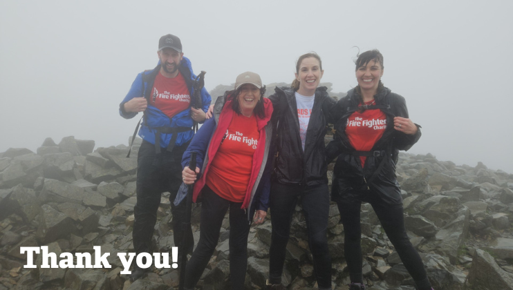 Four people stand smiling on a rocky, foggy summit, wearing rain jackets over red