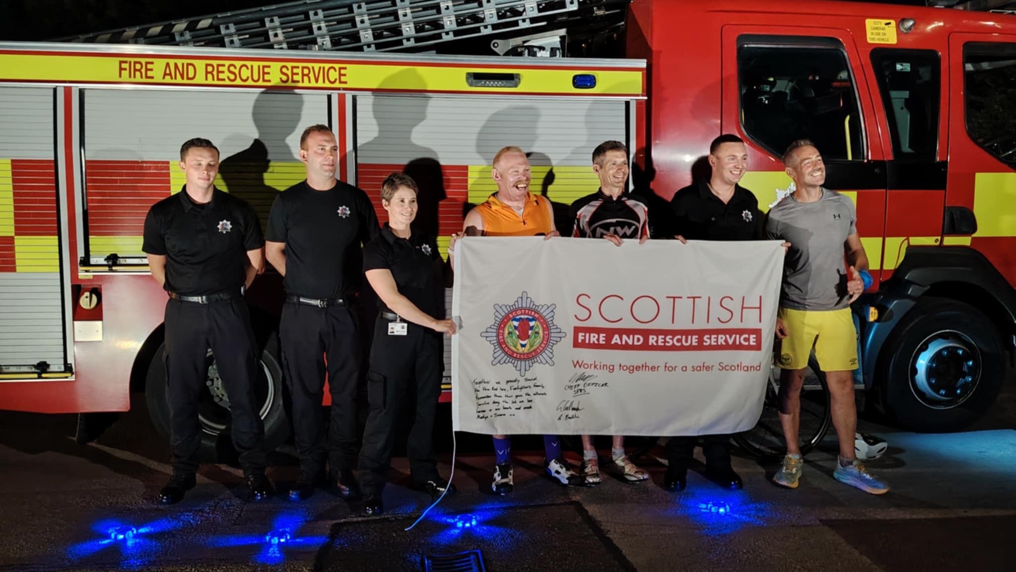 A group of seven people stand in front of a Scottish Fire and Rescue Service fire engine, holding a large banner with the service’s logo and slogan, smiling for the photo at night.