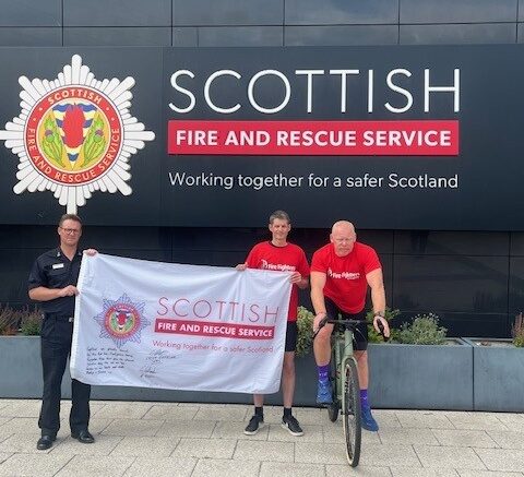 Three people stand outside the Scottish Fire and Rescue Service building. Two hold a banner, one is on a bicycle, and all wear red or black shirts. The building's logo and slogan appear on the wall behind them.