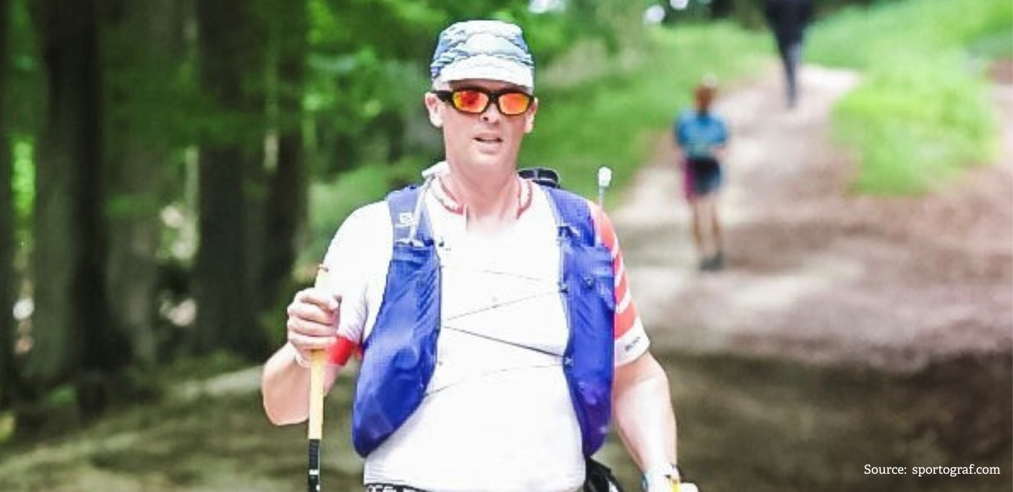 A man wearing sunglasses, a cap, and a hydration vest is trail running through a forested area, holding trekking poles. Another runner is visible on the path in the background.