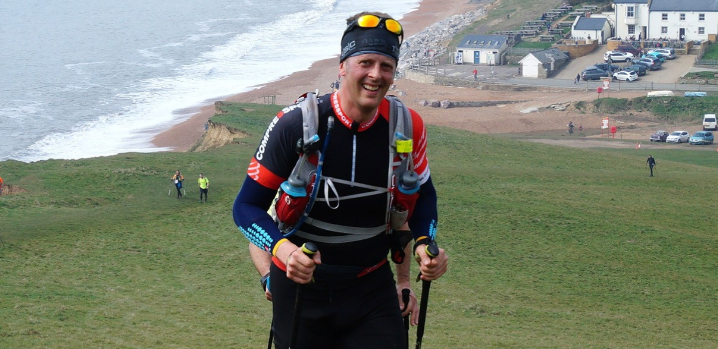 A smiling man wearing athletic gear and sunglasses hikes uphill with trekking poles near a coastal cliff, with the sea, grassy slope, and buildings visible in the background.