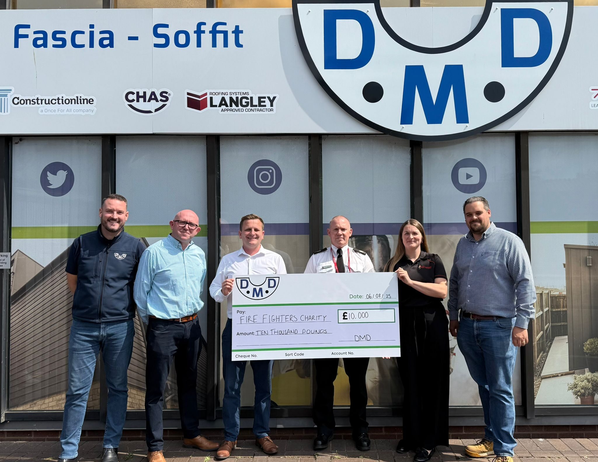 Six people stand smiling outside a DMD building, holding a large ceremonial check for £10,000 made out to the Fire Fighters Charity. The check is dated 24/04/23, and the group appears happy and celebratory.