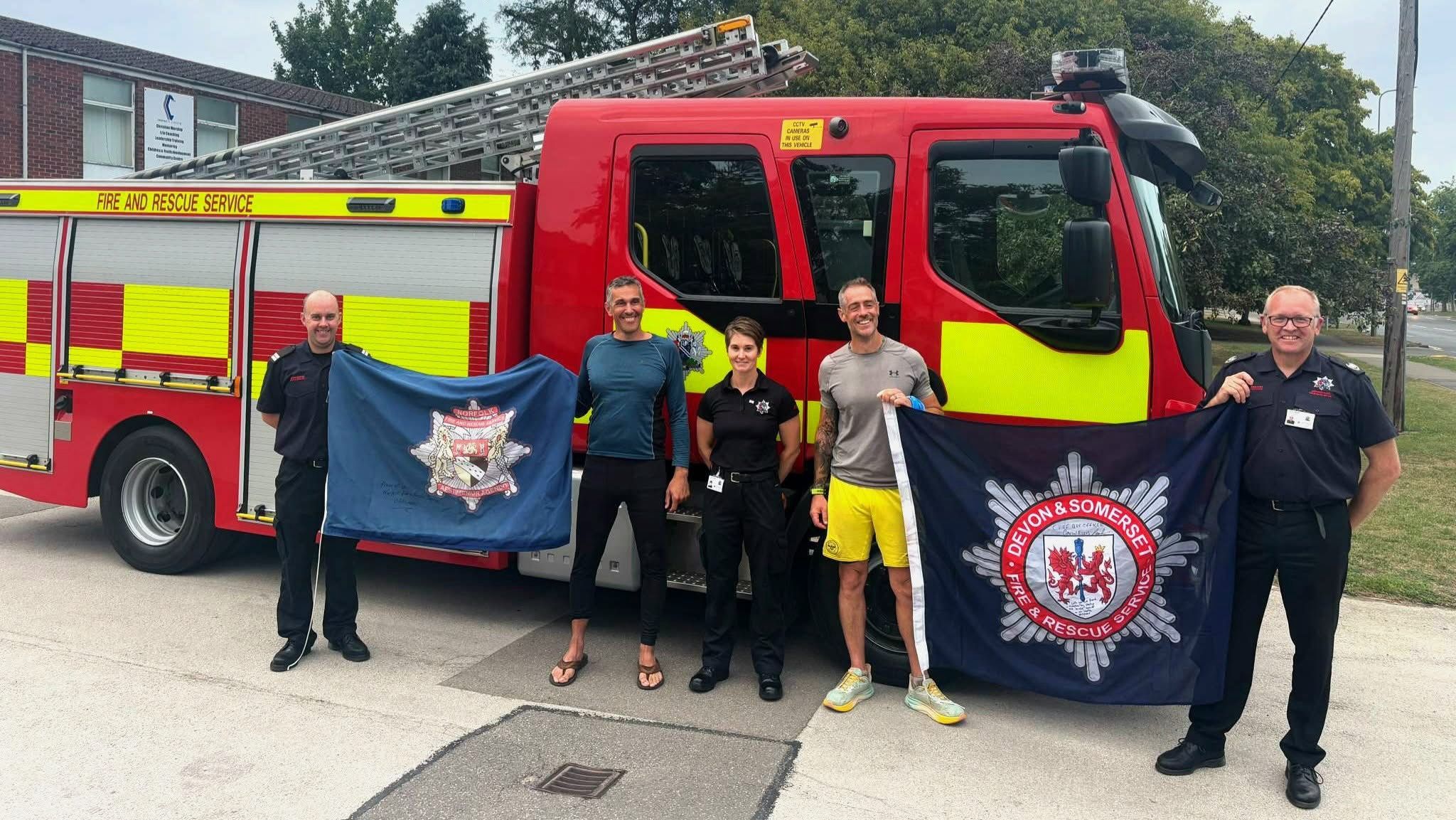 Five people stand smiling in front of a red and yellow fire truck; two hold dark blue flags with fire department logos while the others wear uniforms. Trees and a building are visible in the background.