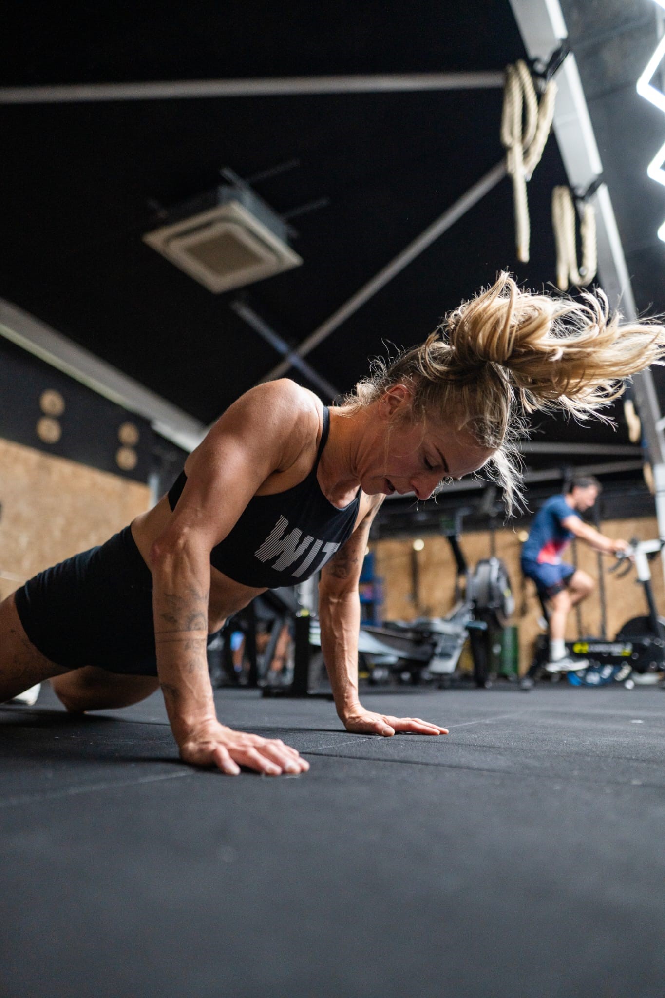 A woman in athletic wear performs a push-up on a gym floor, with her hair in motion. Gym equipment and another person working out are visible in the background.