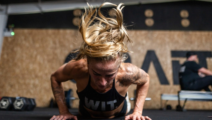 A woman with blond hair, wearing a sports bra labeled “WIT,” does a push-up in a gym, her hair flying upward and her expression focused. Gym equipment and another person are blurred in the background.