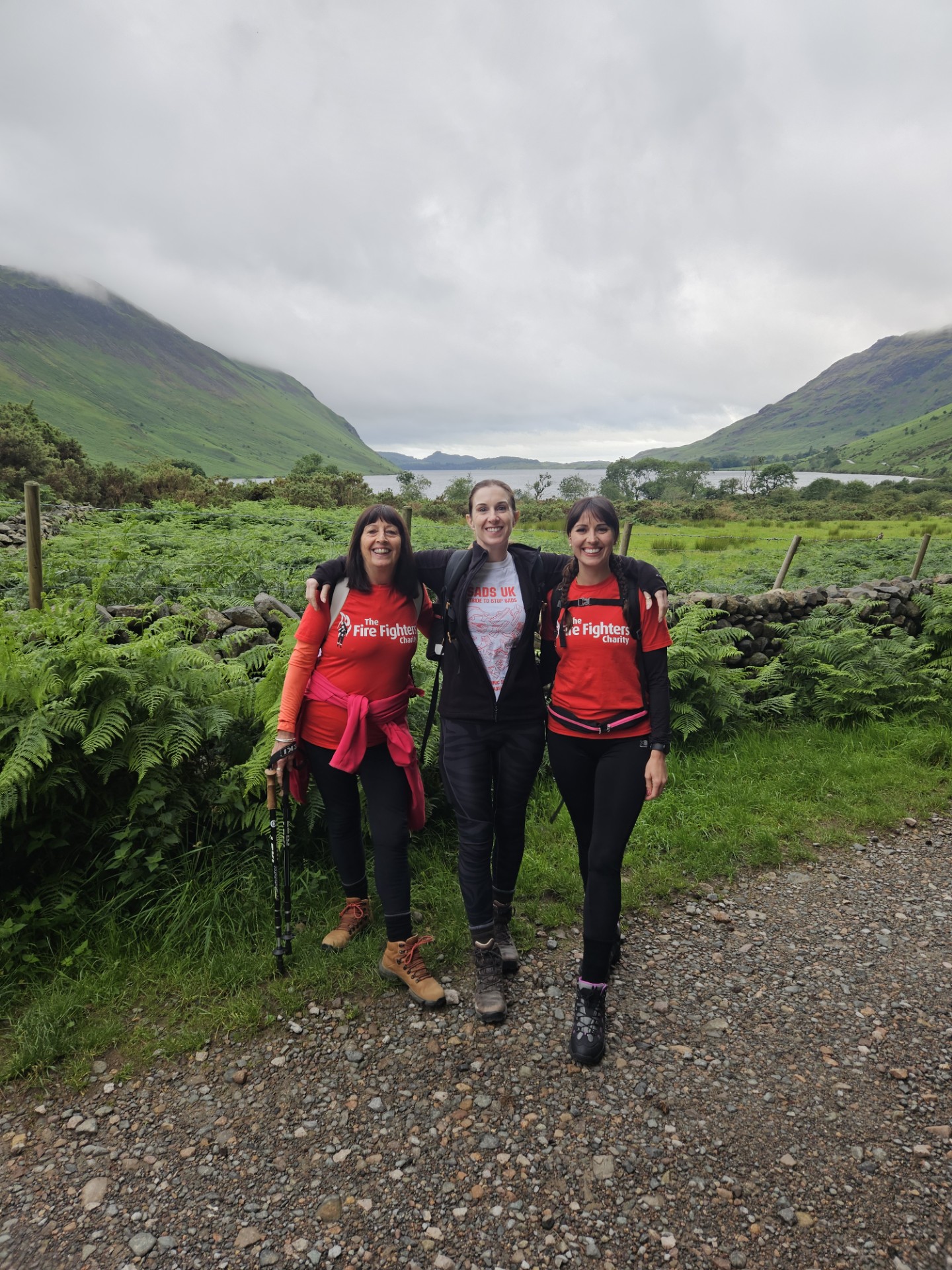 Three women in hiking gear stand smiling on a gravel path in a lush green valley with mountains and cloudy skies in the background. Two wear red "Fire Fighters" shirts, and one wears a white shirt.
