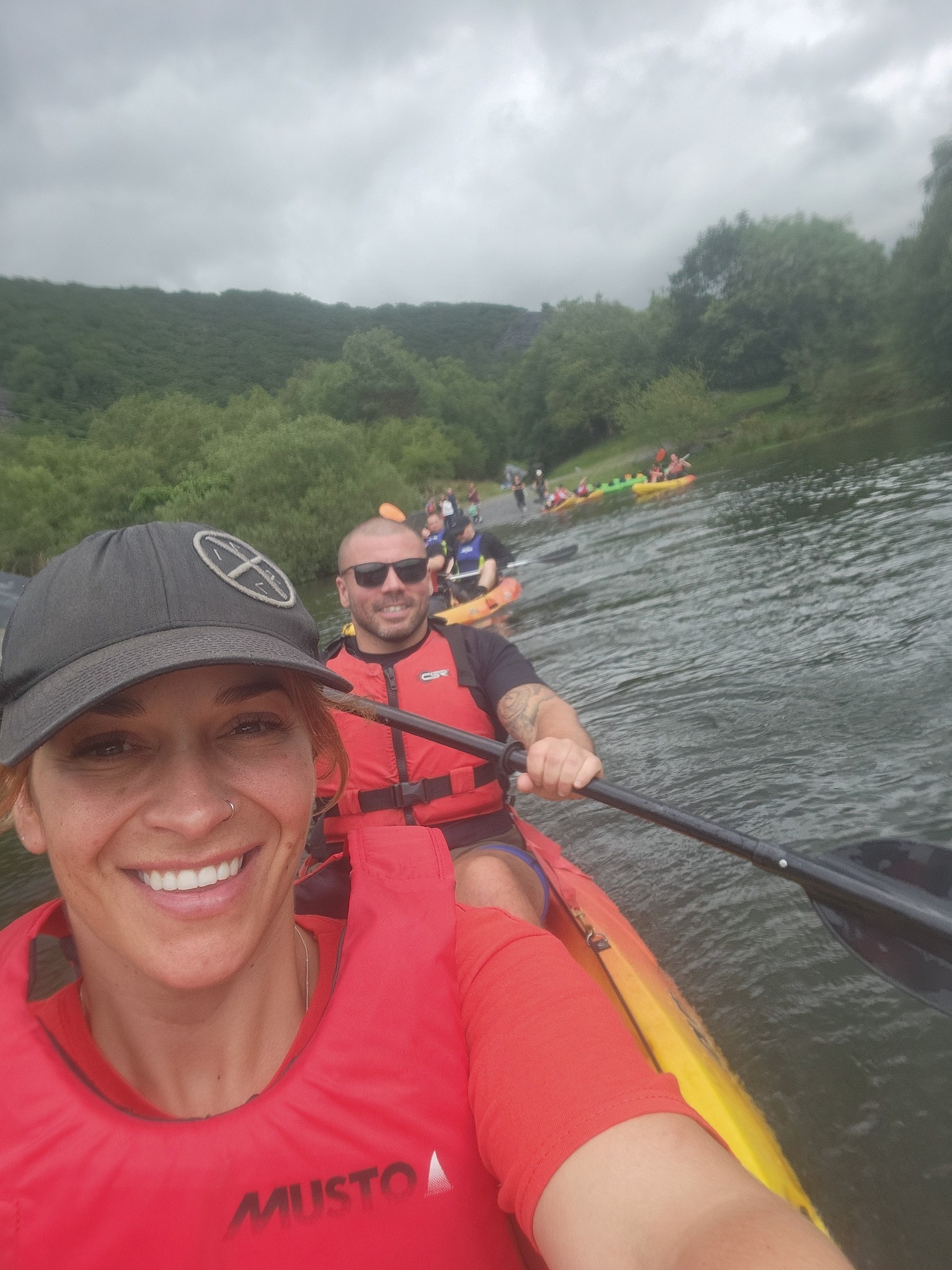 A smiling woman in a red life vest and black cap takes a selfie while kayaking with a man behind her on a river. Other kayakers are visible in the background amid lush greenery and cloudy skies.