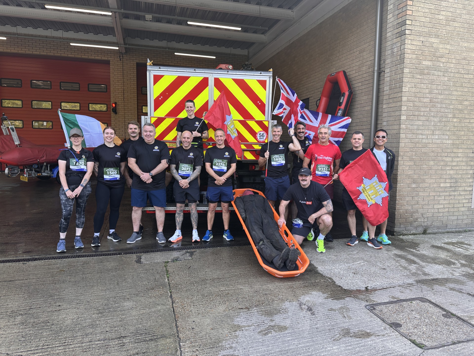 A group of people in athletic gear pose in front of a fire engine in a fire station, holding flags including the UK and Ireland flags, with race bibs on their shirts and a stretcher on the ground in front of them.