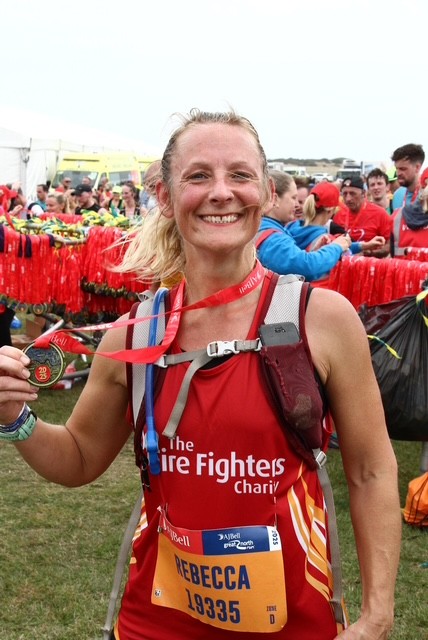 A smiling woman wearing a red running outfit and a race bib labeled "Rebecca 19335" holds up a medal. Behind her, other people and a rack of medals are visible at an outdoor event.
