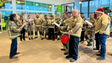 A group of firefighters in uniform, one wearing a Santa hat, stand in a semi-circle inside a building, singing from sheets of paper as one person leads them. Another firefighter holds a red donation bucket.