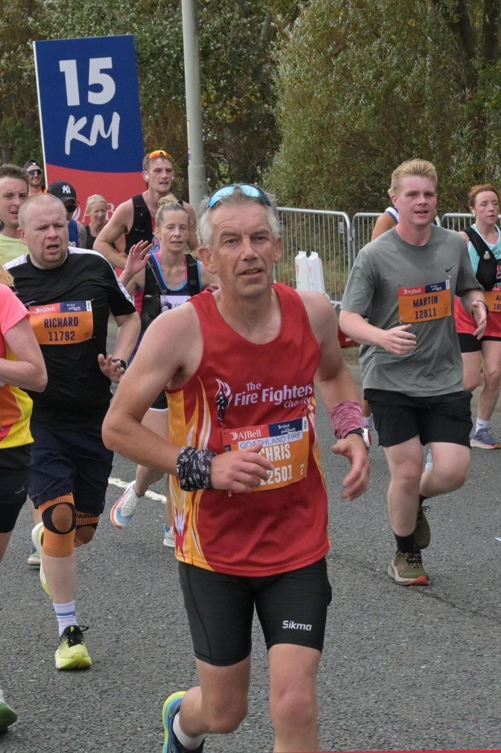 A group of runners participate in a race, passing a "15 KM" marker. The runner in front wears a red "Fire Fighters" vest and a race bib labeled "Chris." Other runners are visible behind him. Trees and barriers line the road.