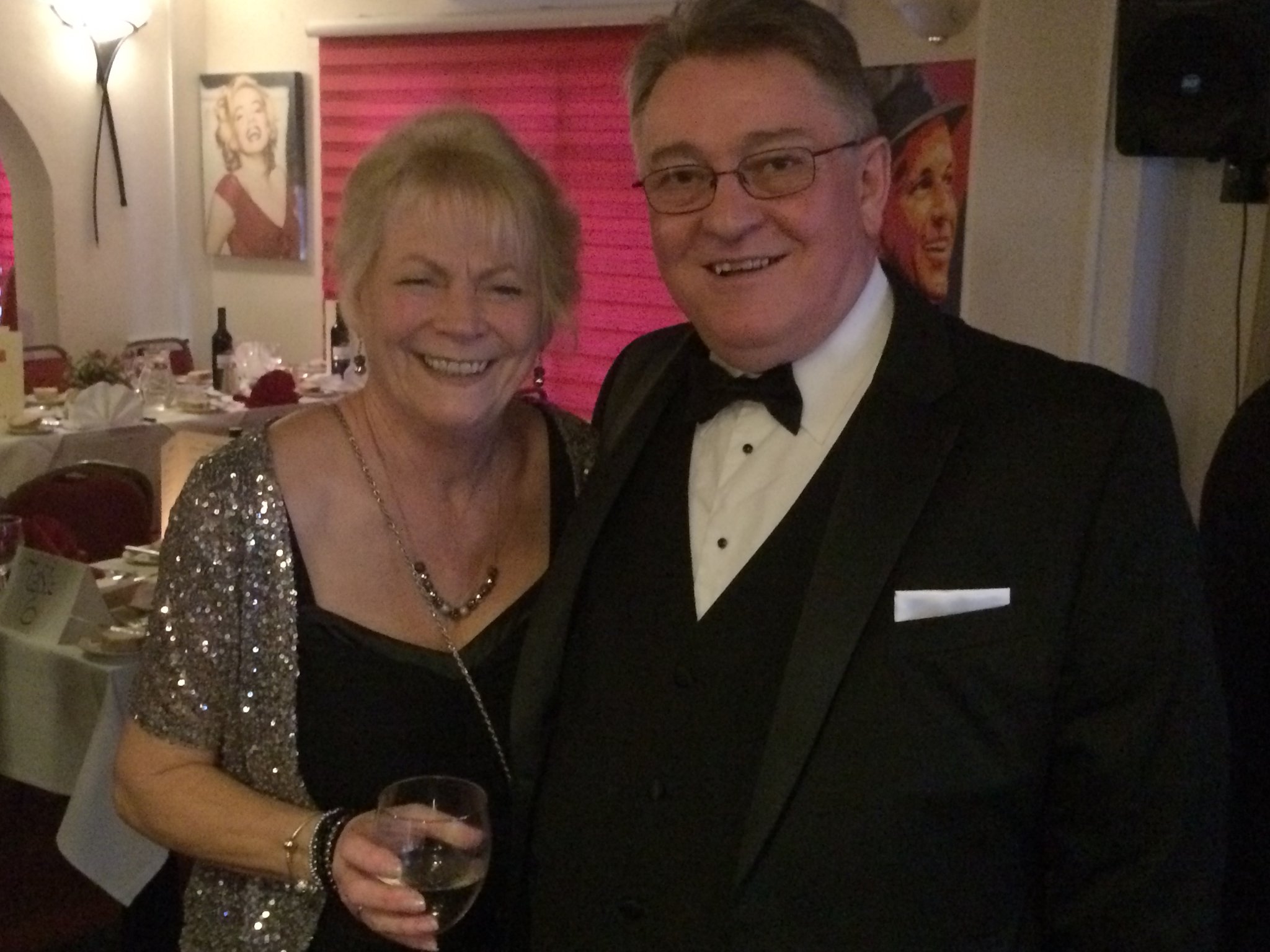A smiling woman in a sparkly jacket and a man in a tuxedo pose together at a formal event. The woman holds a glass, and classic celebrity portraits hang on the wall behind them.