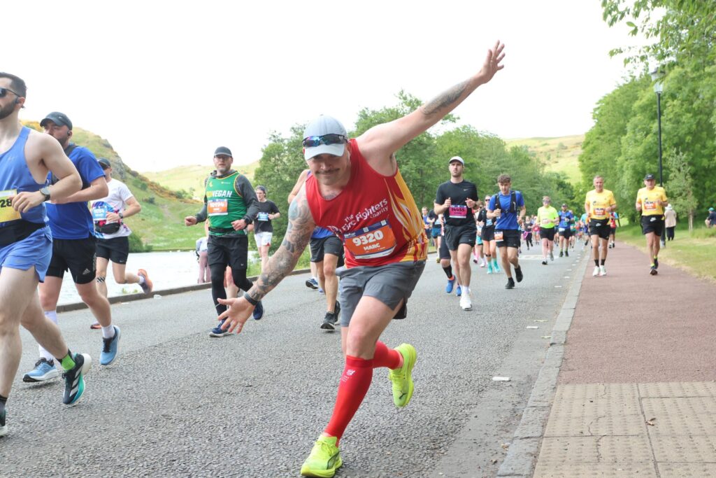 A smiling marathon runner in a red vest and neon shoes poses with arms outstretched toward the camera while other runners continue along a road bordered by greenery.