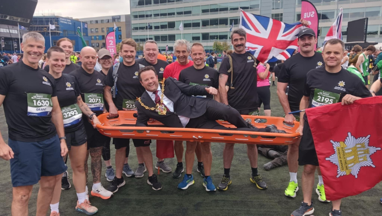 A group of runners pose together outdoors, smiling. They hold an orange stretcher with a man in a suit and mayoral chain lying on it, while others hold UK flags and a red flag with a crest.