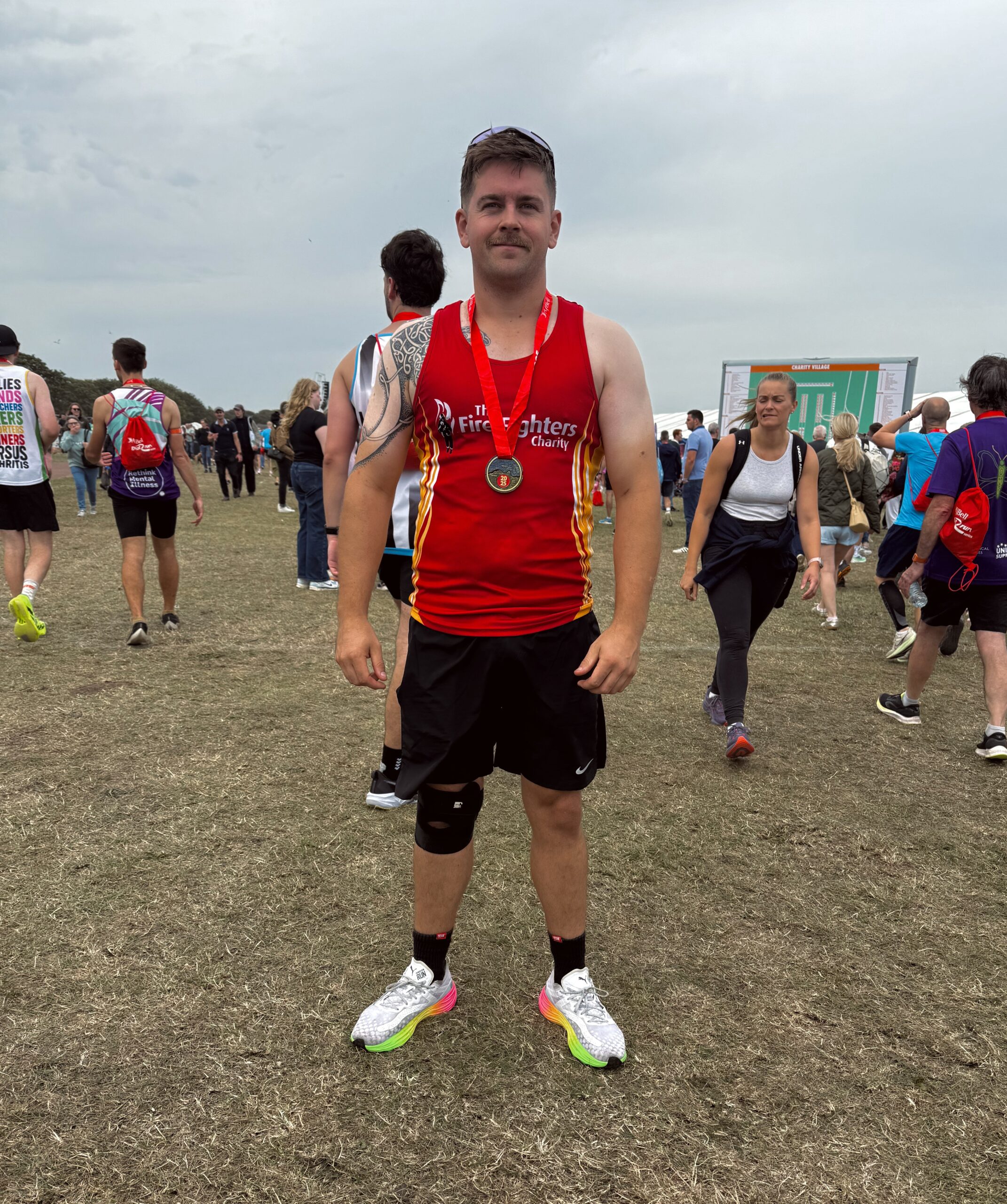 A man wearing a red "Fire Fighters" running vest and black shorts stands on grass with a race medal around his neck. People are walking in the background at an outdoor event on a cloudy day.