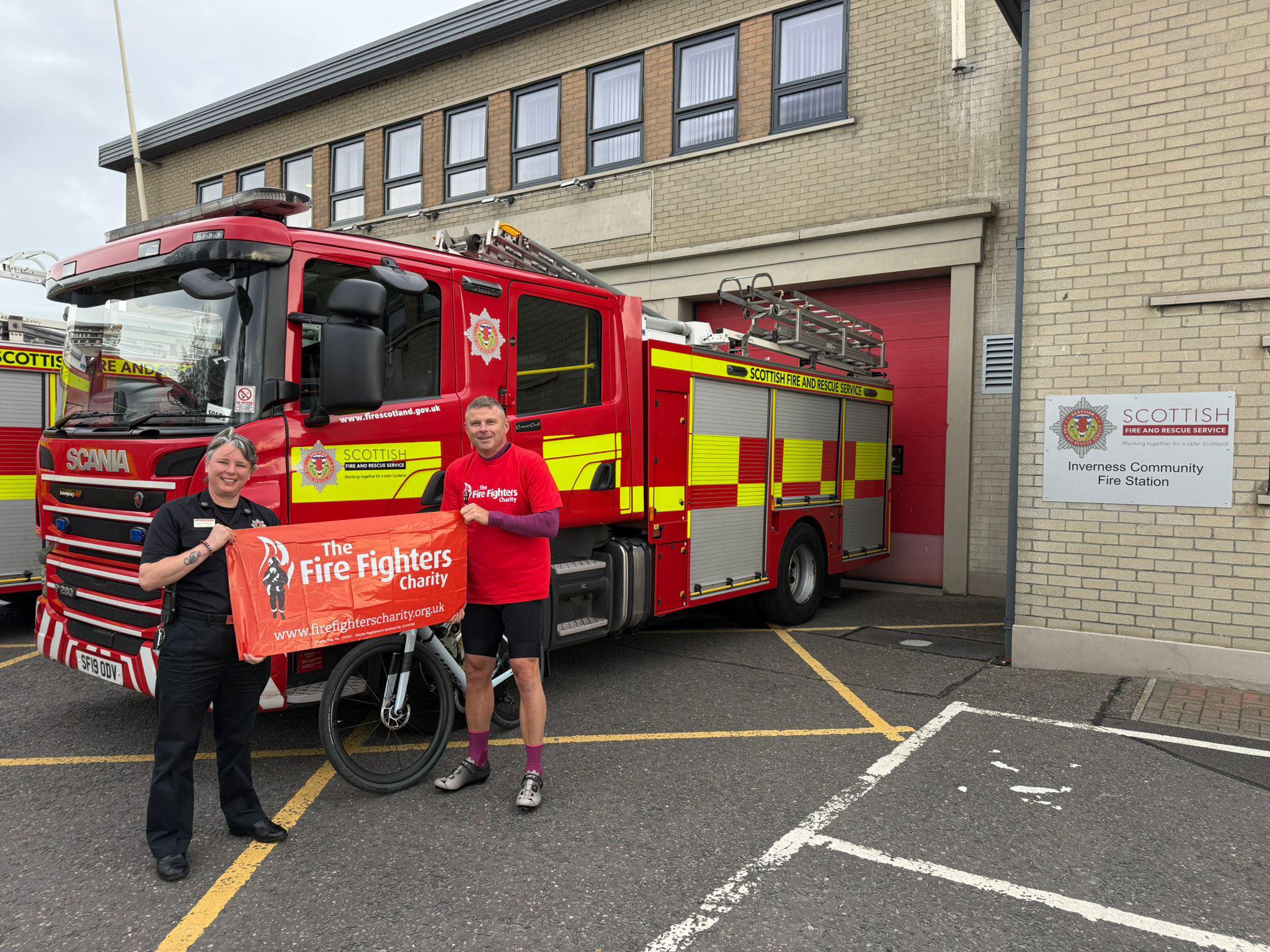 Two people stand in front of a fire engine outside Inverness Community Fire Station, holding an orange banner for The Fire Fighters Charity. The fire engine is parked next to the station’s sign and red garage door.