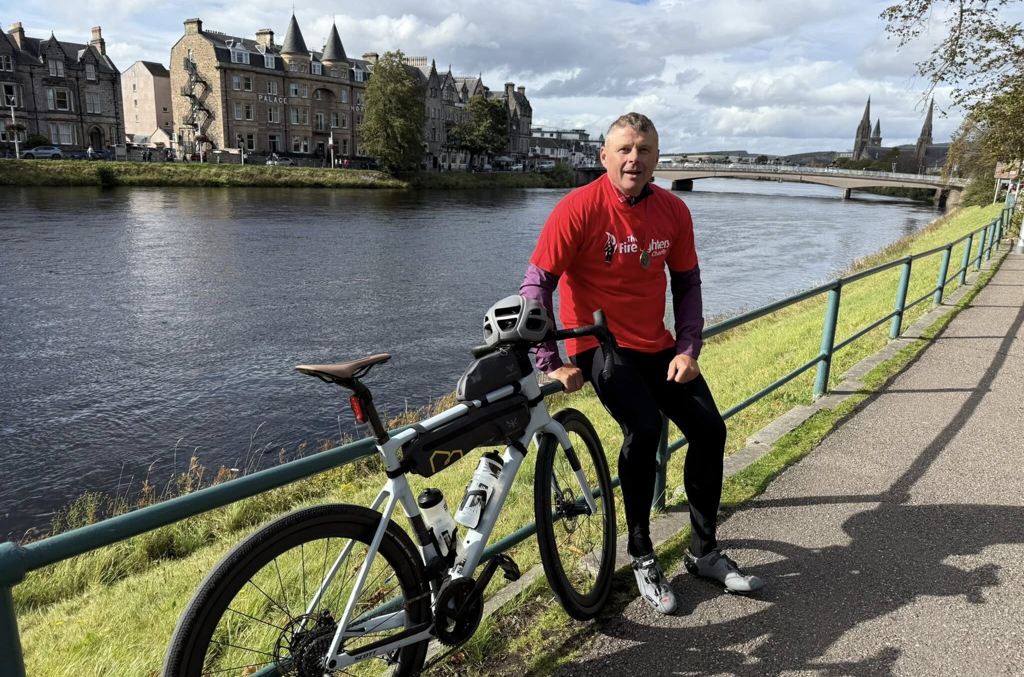 A man in a red shirt sits on a bicycle by a riverside path, with historic buildings and a stone bridge visible across the water under a partly cloudy sky.