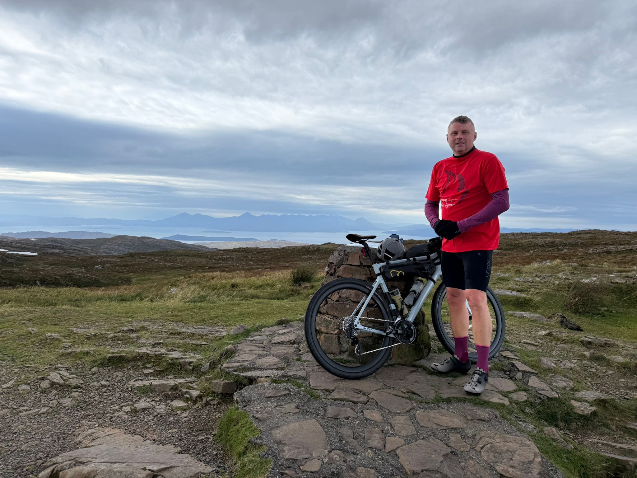 A man in a red shirt and cycling gear stands beside a loaded touring bike on a rocky path, with rolling hills and a cloudy sky in the background.