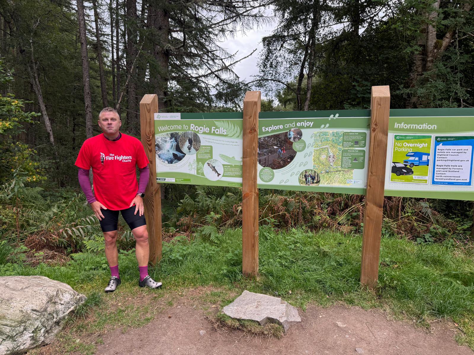 A man in a red shirt and cycling gear stands by three large informational signs at the Rogie Falls forest entrance, surrounded by trees and greenery.