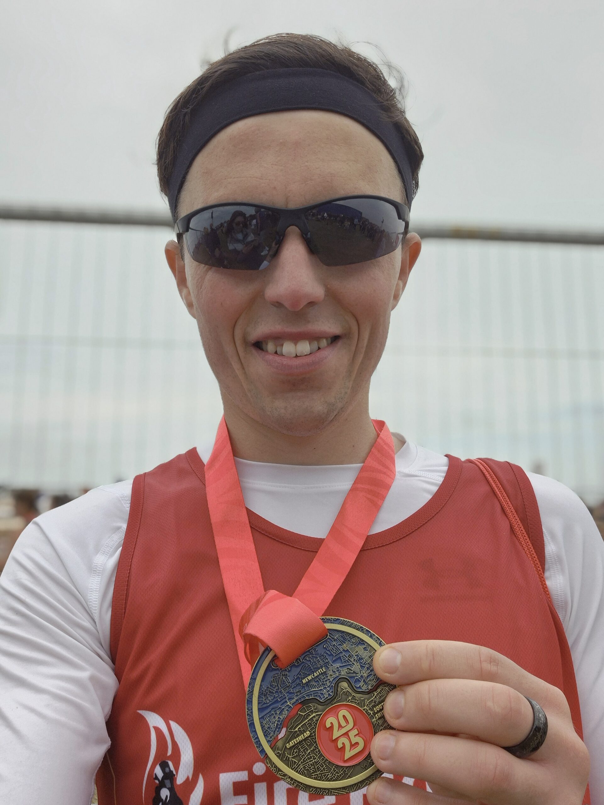 A smiling runner wearing sunglasses and a red headband holds up a medal with a red ribbon. The medal features a scenic design and the numbers "22 25." The runner is dressed in a red and white athletic top.