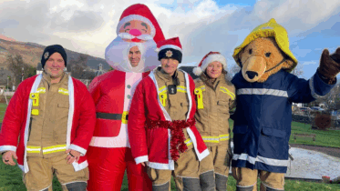 Five people stand outdoors: three in firefighter uniforms, one in an inflatable Santa costume, and one in a bear mascot suit wearing a firefighter coat and hat. The group smiles, with a scenic hillside in the background.