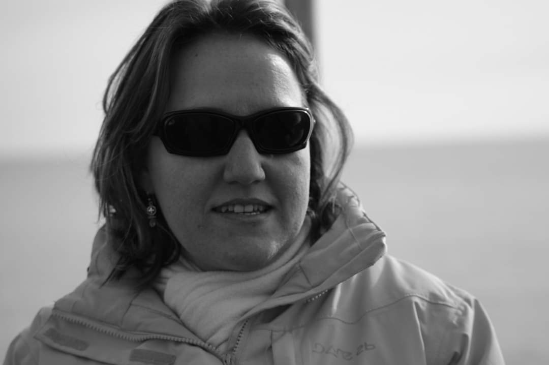 A woman wearing dark sunglasses, a light jacket, and a scarf, stands outdoors. The image is in black and white, with a blurred background that suggests a calm, open area, possibly near water.