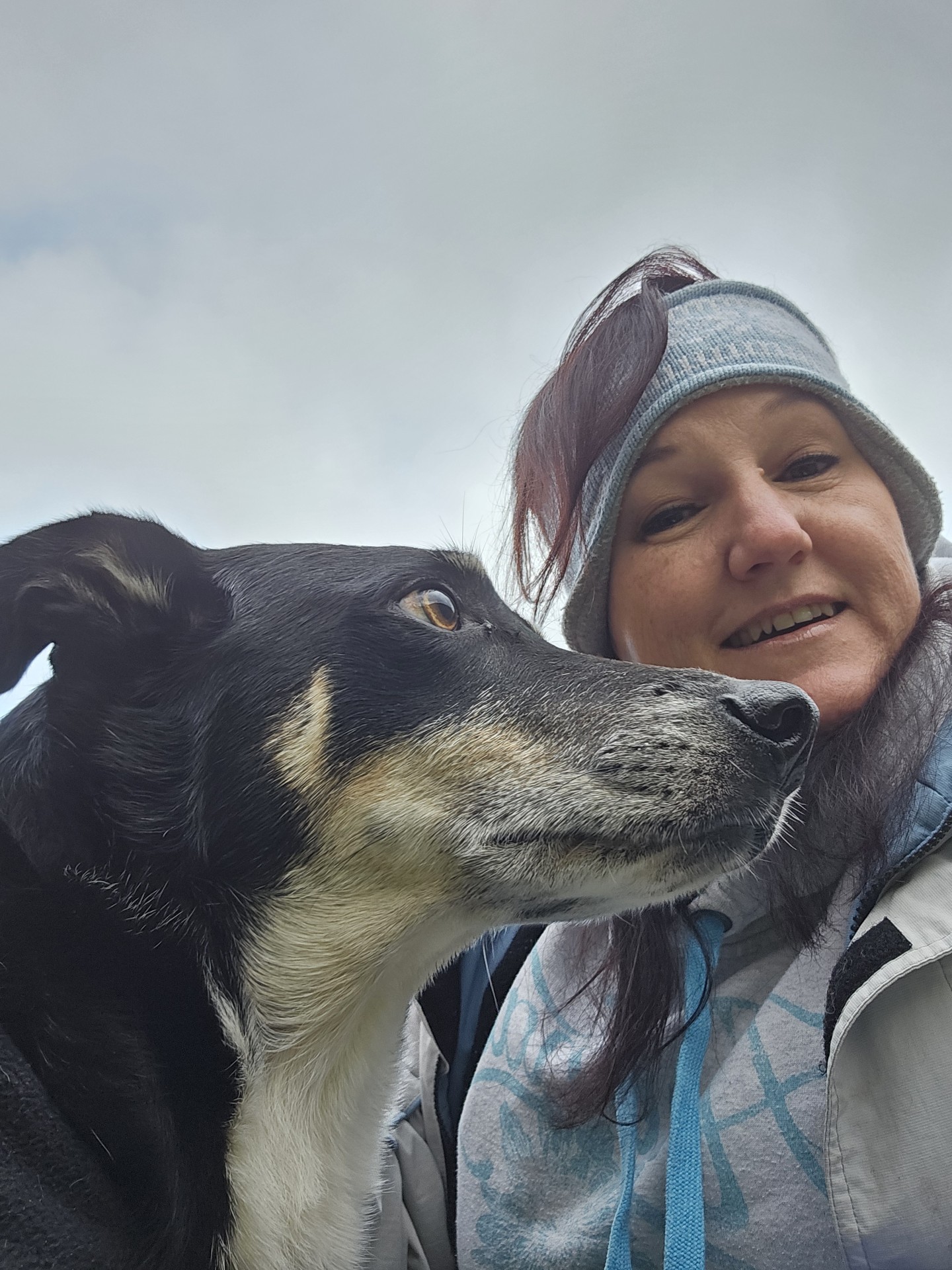 A woman wearing a headband and jacket smiles at the camera while posing outdoors with a black and tan dog under a cloudy sky.