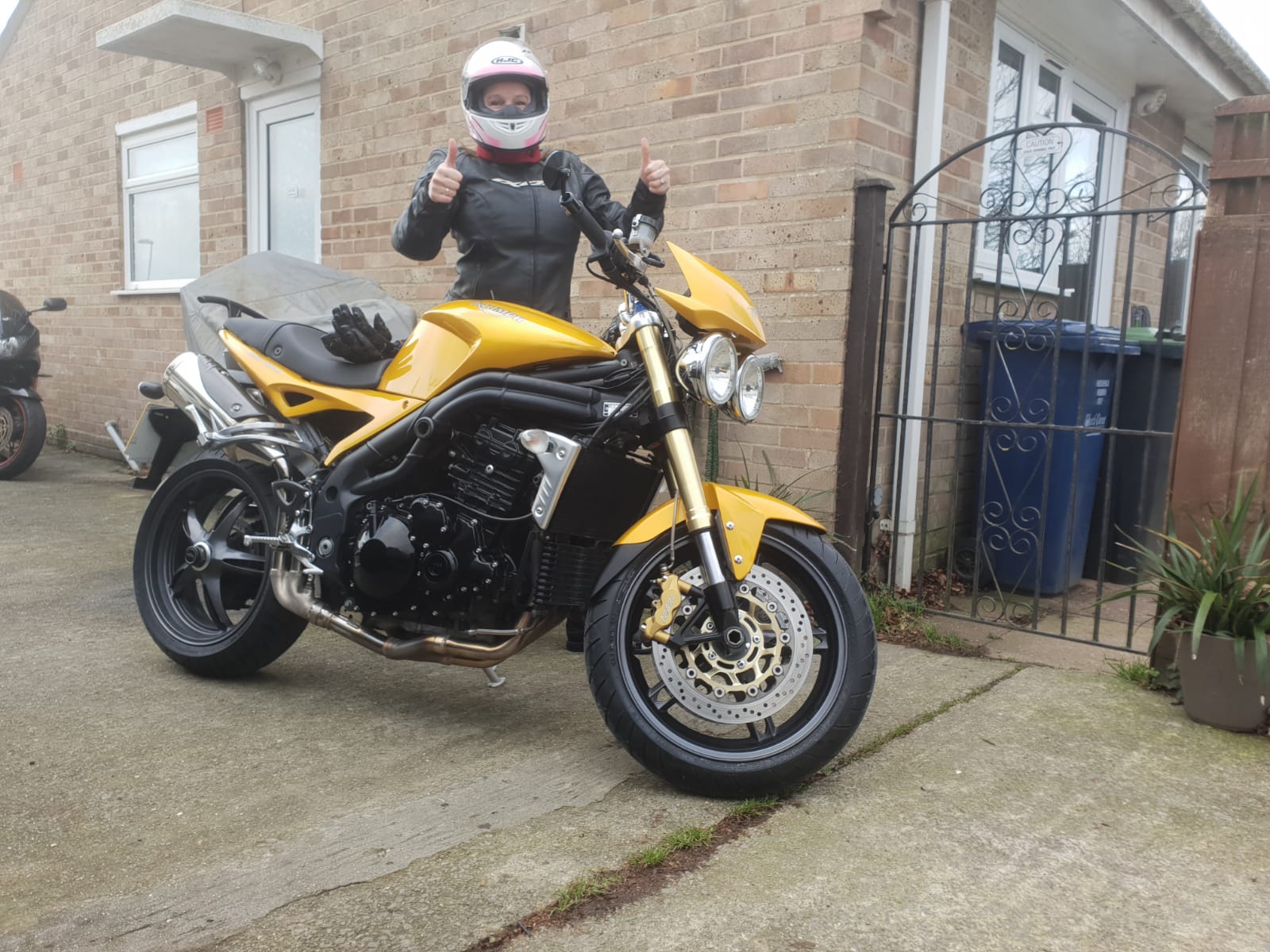 A person wearing a helmet and motorcycle gear stands behind a yellow motorcycle, giving two thumbs up, in a residential driveway near a brick house and a metal gate.