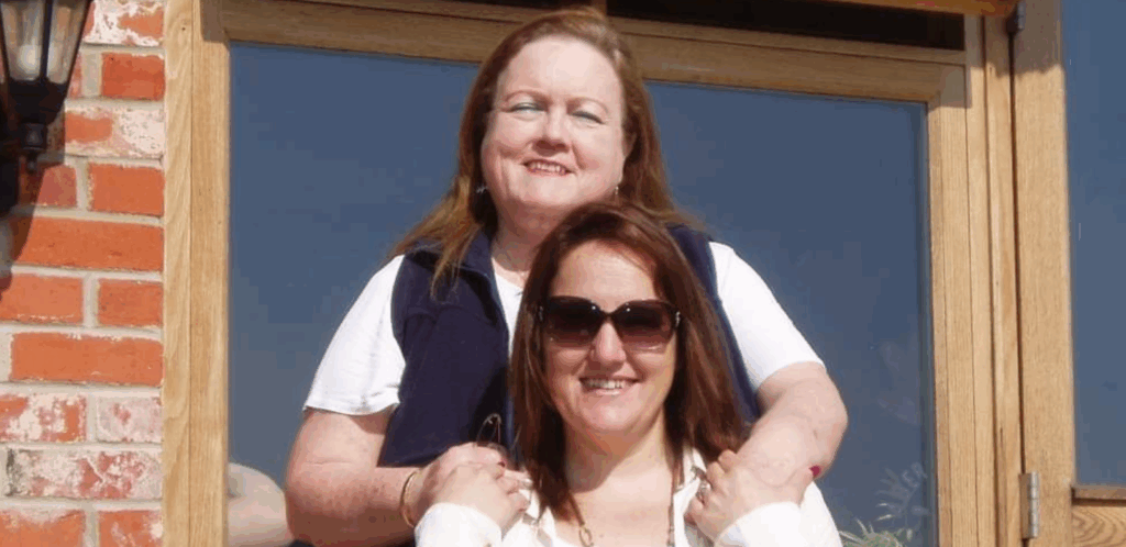 Two women smiling outdoors in front of a wooden-framed window; the woman in back has her hands on the shoulders of the woman in front, who is wearing sunglasses. Sunlight is shining on both of them.