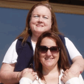 Two women smiling outdoors in front of a wooden-framed window; the woman in back has her hands on the shoulders of the woman in front, who is wearing sunglasses. Sunlight is shining on both of them.