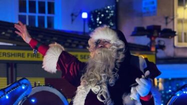 Santa Claus dressed in a red suit and white gloves waves cheerfully while sitting in a festive sleigh at night, with blue lights and a building in the background.