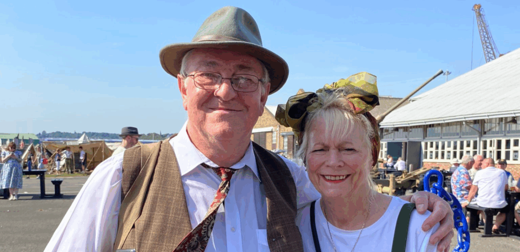 An older man and woman, both smiling and dressed in vintage-style clothing, stand outside on a sunny day at an event with tents and people in the background.