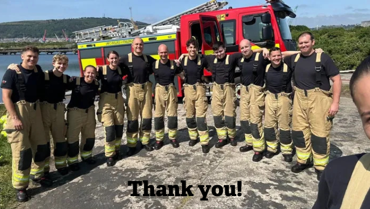 A group of firefighters in uniform stand smiling in front of a fire truck on a sunny day, with
