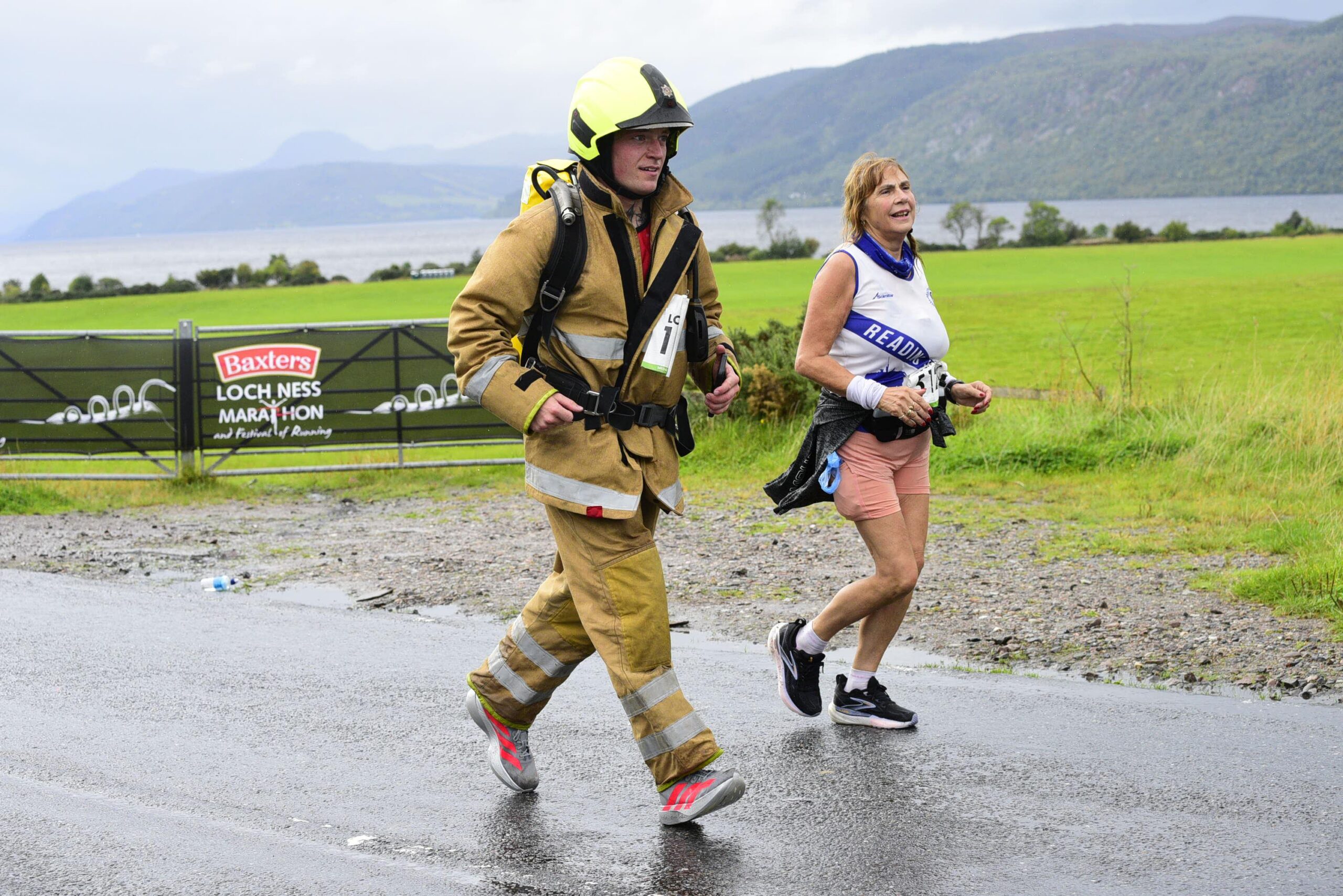 A firefighter in full turnout gear and a runner in athletic clothes running side-by-side on a road near the Loch Ness Marathon banner.