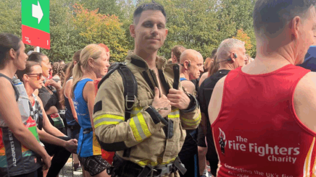 A man in a firefighter uniform stands among marathon runners at the starting line, surrounded by people wearing athletic gear. Trees and a green sign are visible in the background.