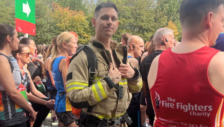 A man in a firefighter uniform stands among marathon runners at the starting line, surrounded by people wearing athletic gear. Trees and a green sign are visible in the background.