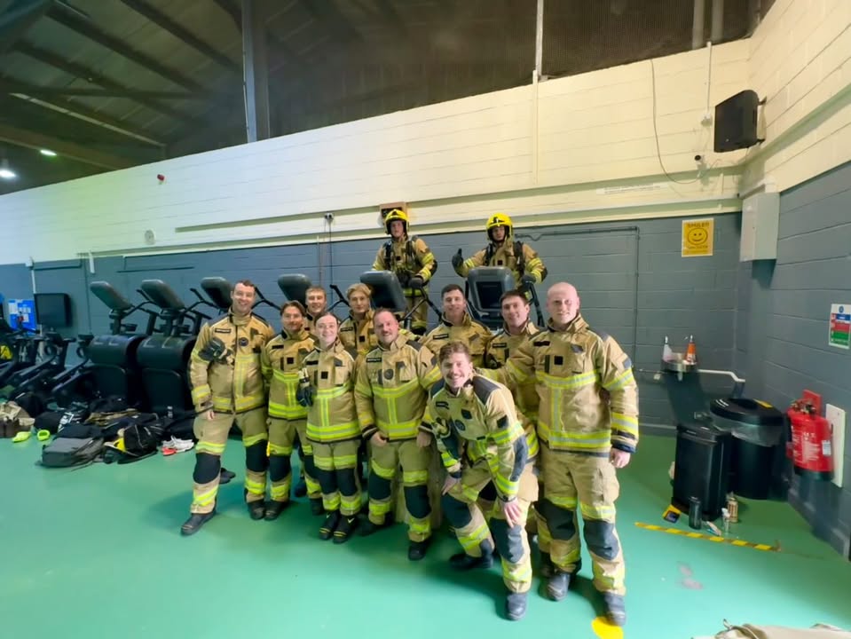 A group of firefighters in full gear pose and smile together in a gym-like indoor setting, with two standing on exercise equipment behind the group.