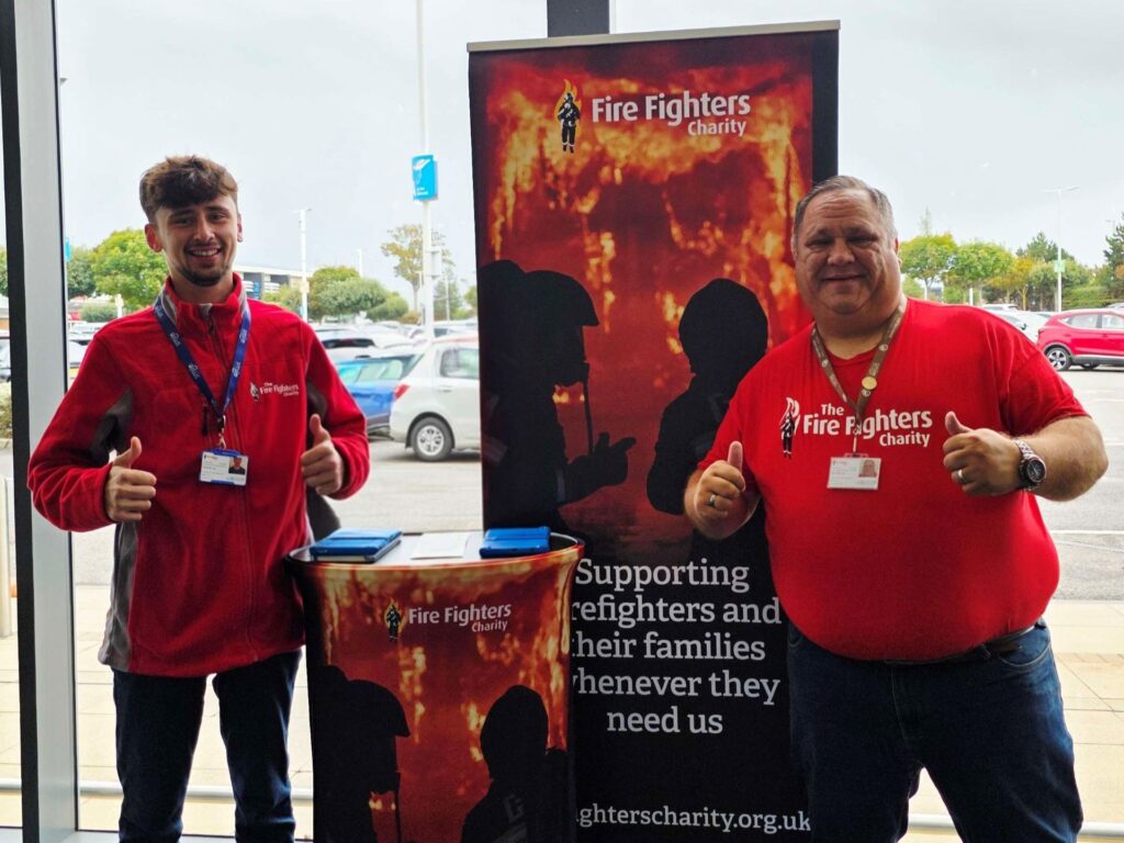 Two men wearing red "Fire Fighters Charity" shirts stand indoors, smiling and giving thumbs up next to a Fire Fighters Charity banner and donation box. The banner reads, "Supporting firefighters and their families whenever they need us.