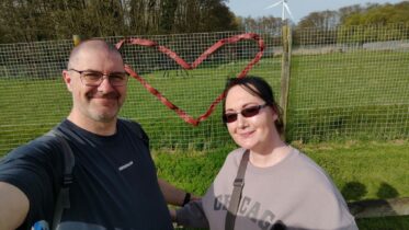 A man and woman smile for a selfie in front of a green field, with a fence behind them decorated with a red heart shape. Trees and a wind turbine are visible in the background under a clear sky.