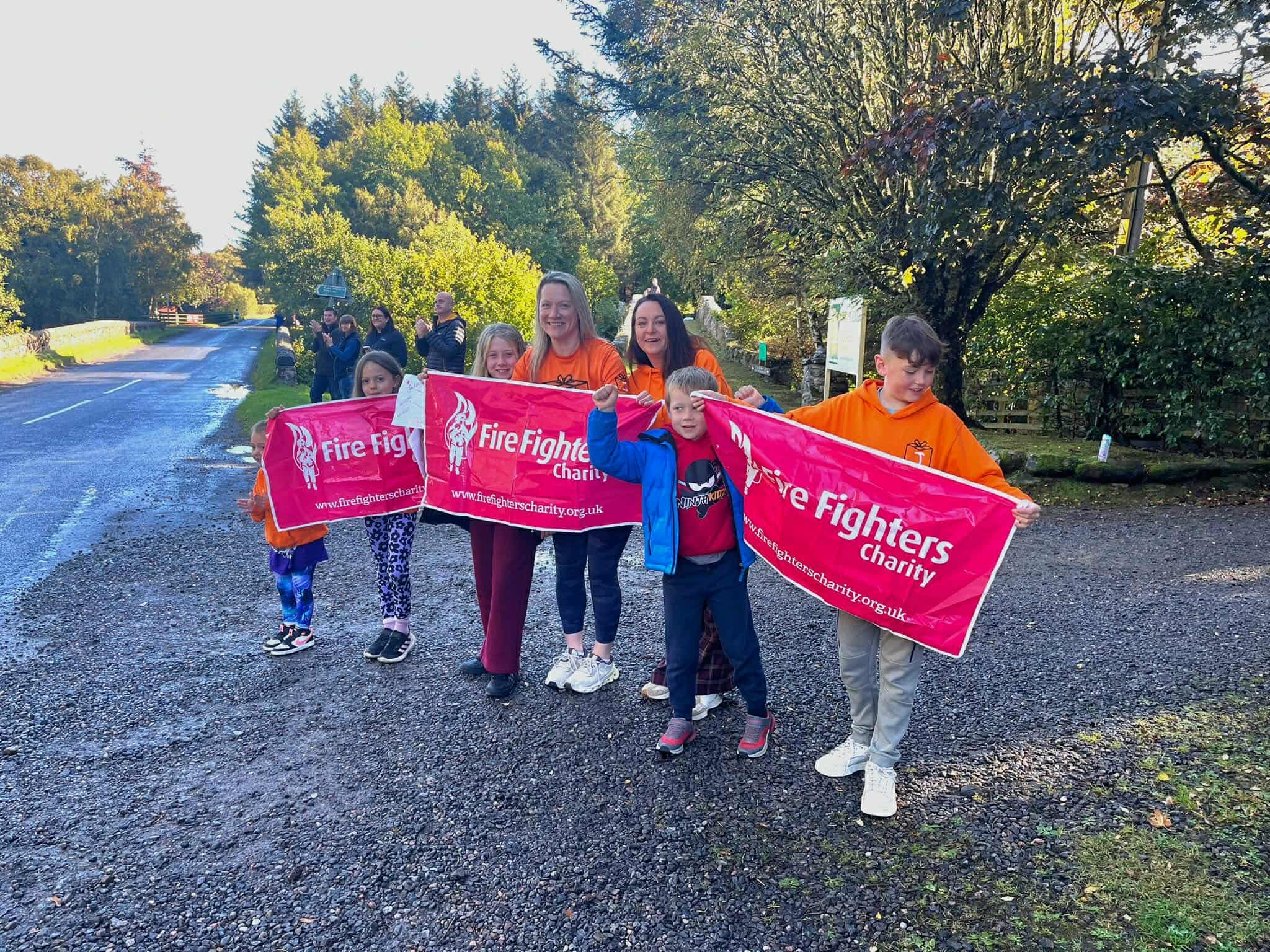 A group of adults and children stand outdoors on a gravel path, smiling and holding two bright red “Fire Fighters Charity” banners. Some people in the background are also present along a road lined with trees and greenery.