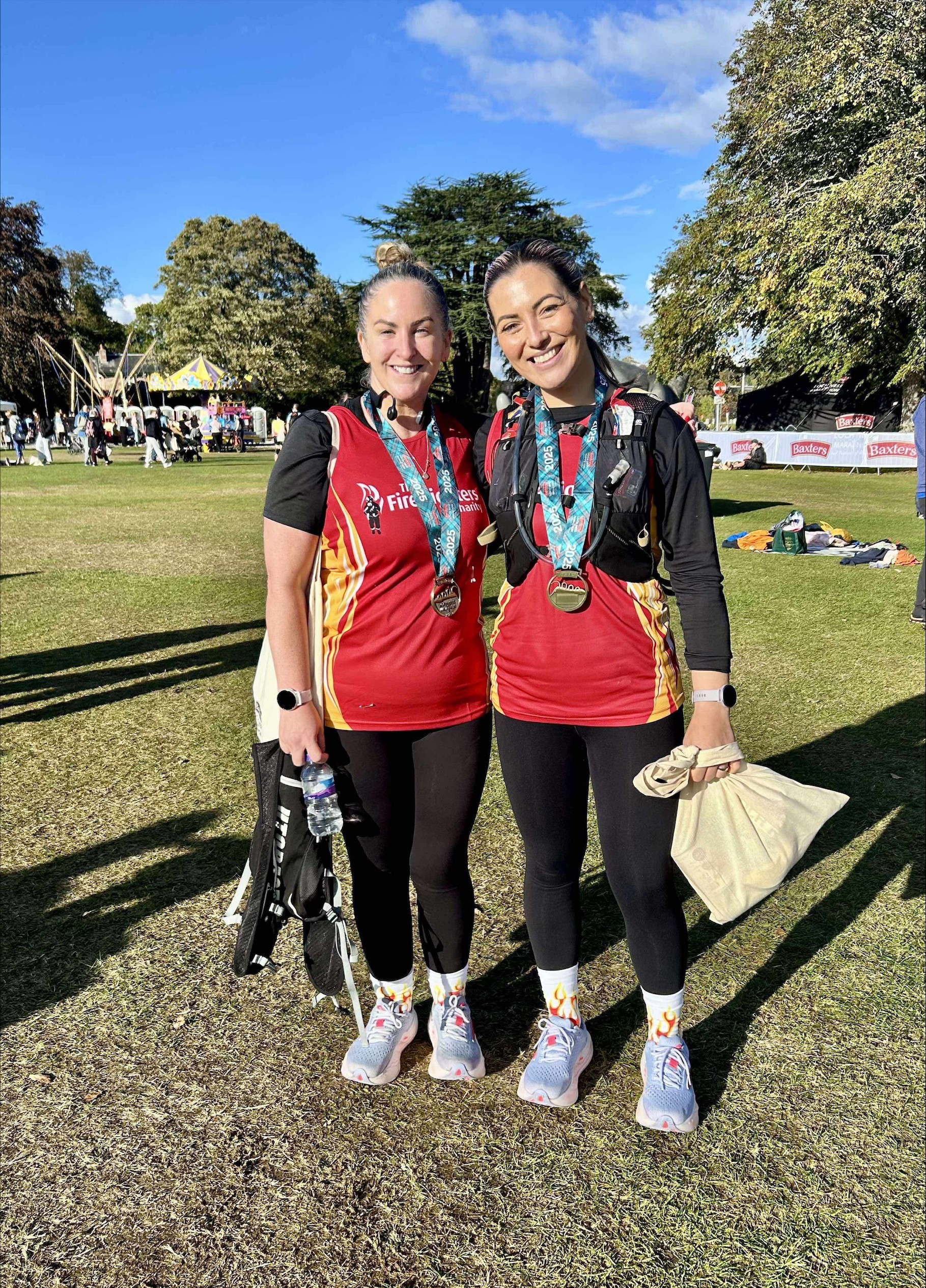 Two women stand side by side on grass, smiling at the camera. Both wear red running shirts, black leggings, and medals around their necks, suggesting they’ve completed a race. People and trees are visible in the background.