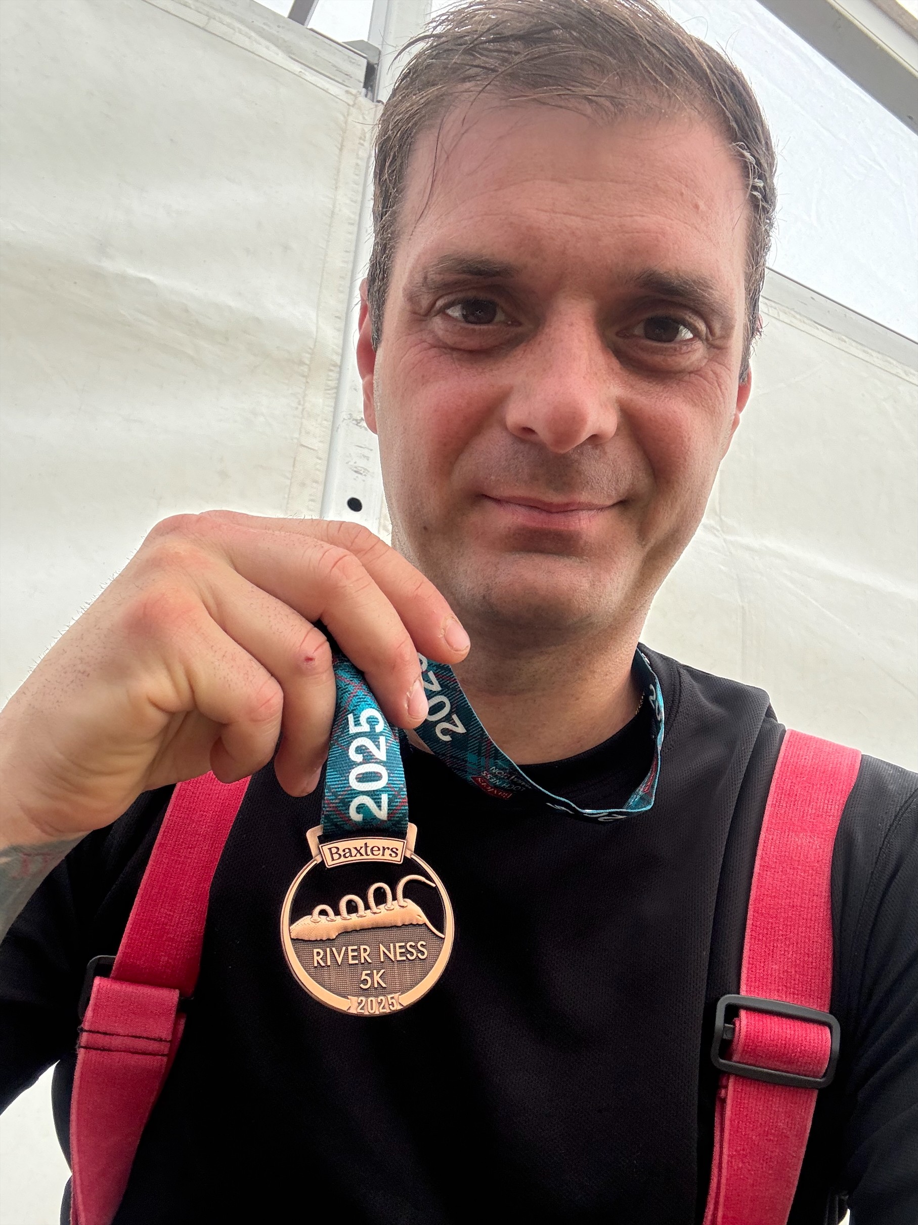 A man wearing a black shirt and red suspenders smiles and holds up a River Ness 5K 2023 medal, with a "2023" lanyard visible. He appears sweaty and happy, standing in front of a white background.