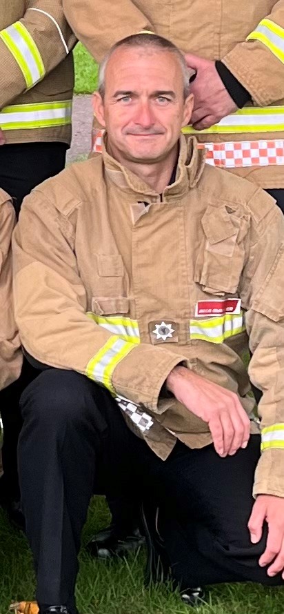 A firefighter kneels on grass, wearing a tan fire-resistant jacket with reflective yellow stripes. He is smiling at the camera, with other firefighters standing nearby, partially visible.