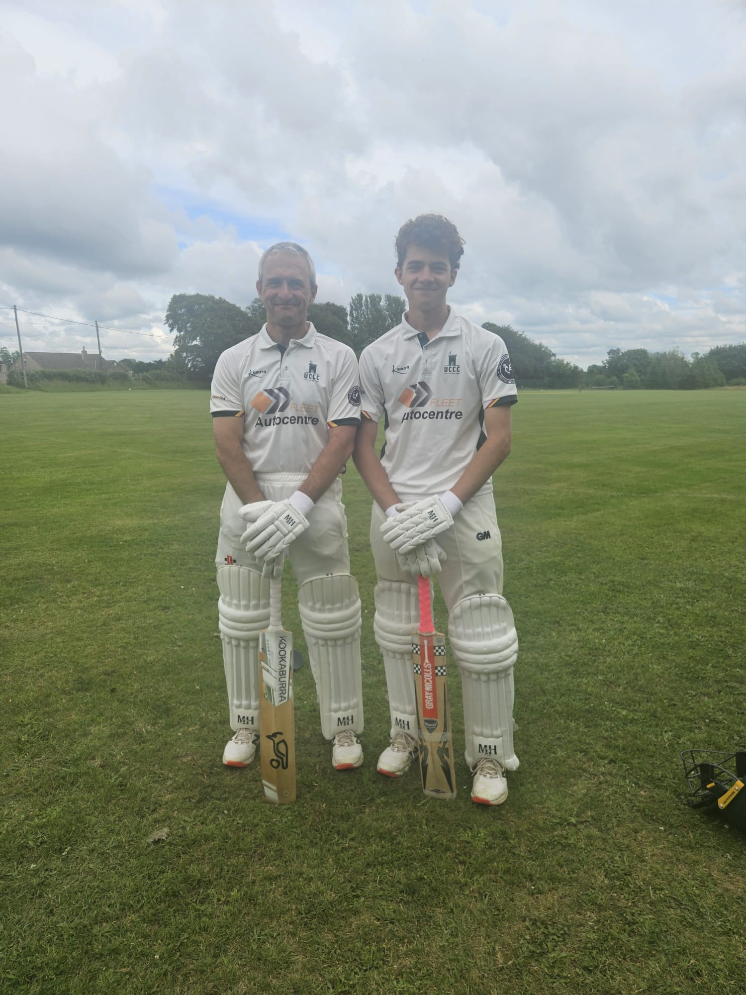 Two cricketers wearing white uniforms and pads stand side by side on a grassy cricket field, each holding a bat. The sky is cloudy, and trees are visible in the background.