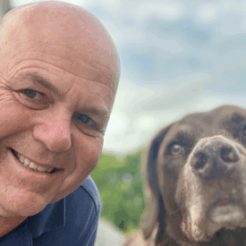 A smiling bald man in a blue shirt takes a selfie outdoors next to a brown dog. The background shows greenery and a cloudy sky.