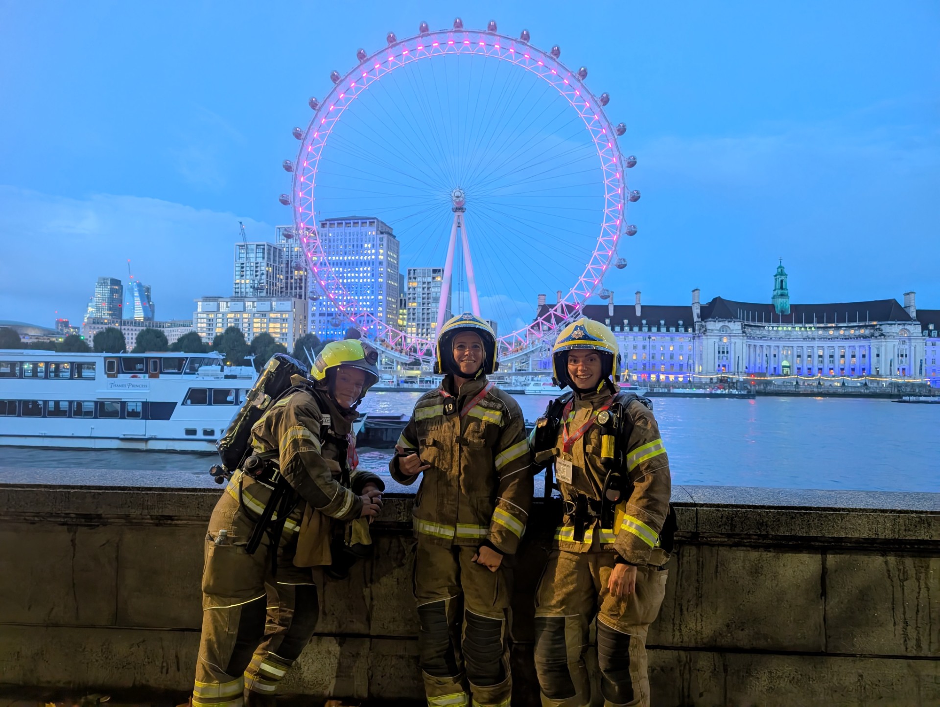 Three firefighters in full gear stand and smile in front of the River Thames with the illuminated London Eye and city buildings in the background during dusk.